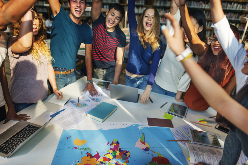 Eine heterogene Schüler:innengruppe hebt begeistert die Hände über einer Weltkarte