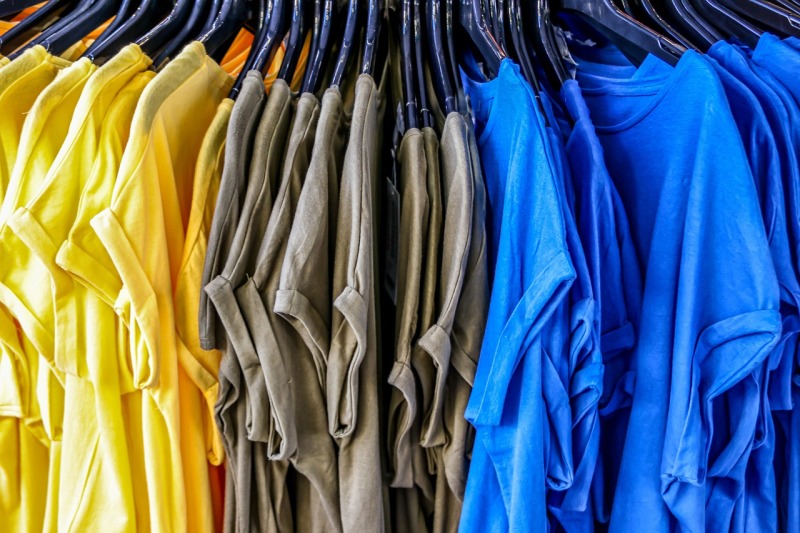 Das Bild zeigt eine Menge gleicher T-Shirts, die auf Plastikbügeln nebeneinander aufgehängt sind. Sie haben die Farben gelb, olivgrün und blau.