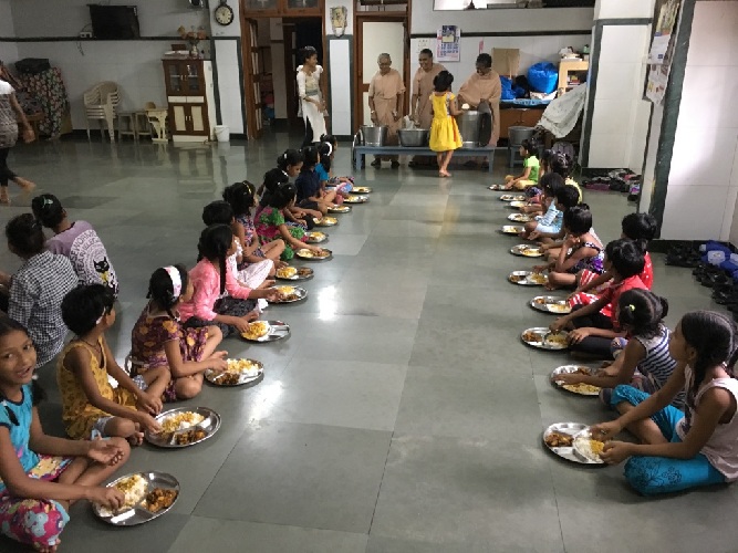 Kinder sitzen im Speisesaal auf dem Boden, vor sich einen Teller mit Essen, im Hintergrund die Essensausgabe