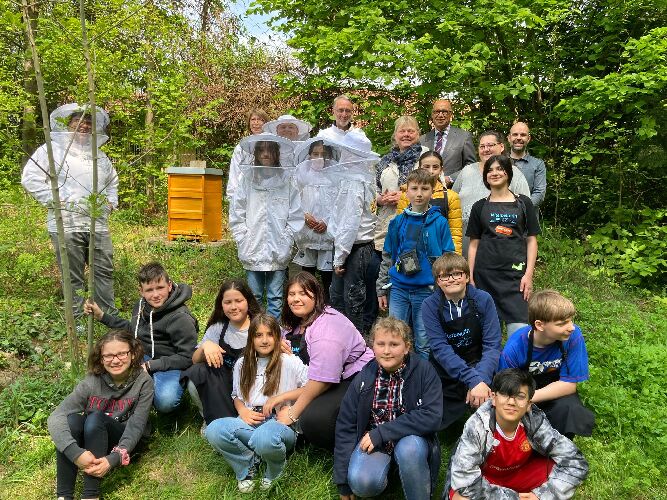 Bild einer Menschengruppe im Schulgarten neben dem neuen Bienenstock bestehend aus Mitgliedern der Schulgarten AG und des Imkers Herr Bergmann, der Fairtrade-Gruppe und der Schulleitung der Friedensschule.