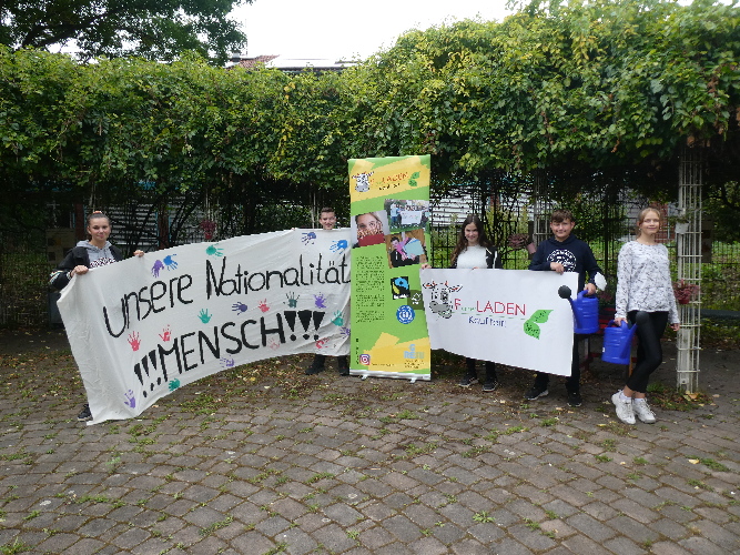 Auf dem Foto präsentieren Schülerinnen und Schüler (von links nach rechts) ein Demonstrationsplakat der Schülerdemonstration gegen Rassismus, zwei Werbeplakate des FLaden (FairerLaden) und für die  AG 
