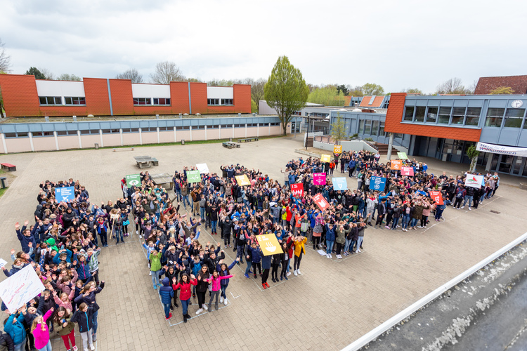 Alle Schüler der Schule stehen auf dem Schulhof in Form der Buchstaben HNGE, dem Namen unserer Schule, Heinz-Nixdorf-Gesamtschule. Dabei halten die Schüler die Bilder aller 17 SDGs nach oben und schauen in die Kamera einer Drohne.