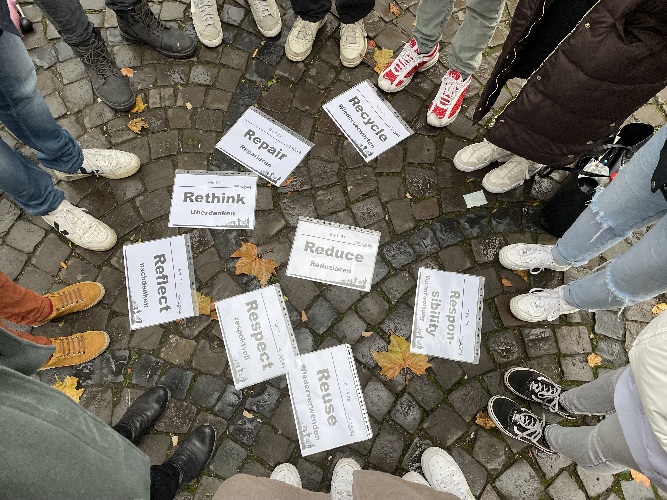 Die Schülerinnen und Schüler sind im Kreis in der Kölner Innenstadt aufgestellt. In der Mitte liegen Prints mit Empfehlungen nachhaltiger Handlungsmöglichkeiten eines jeden Einzelnen.