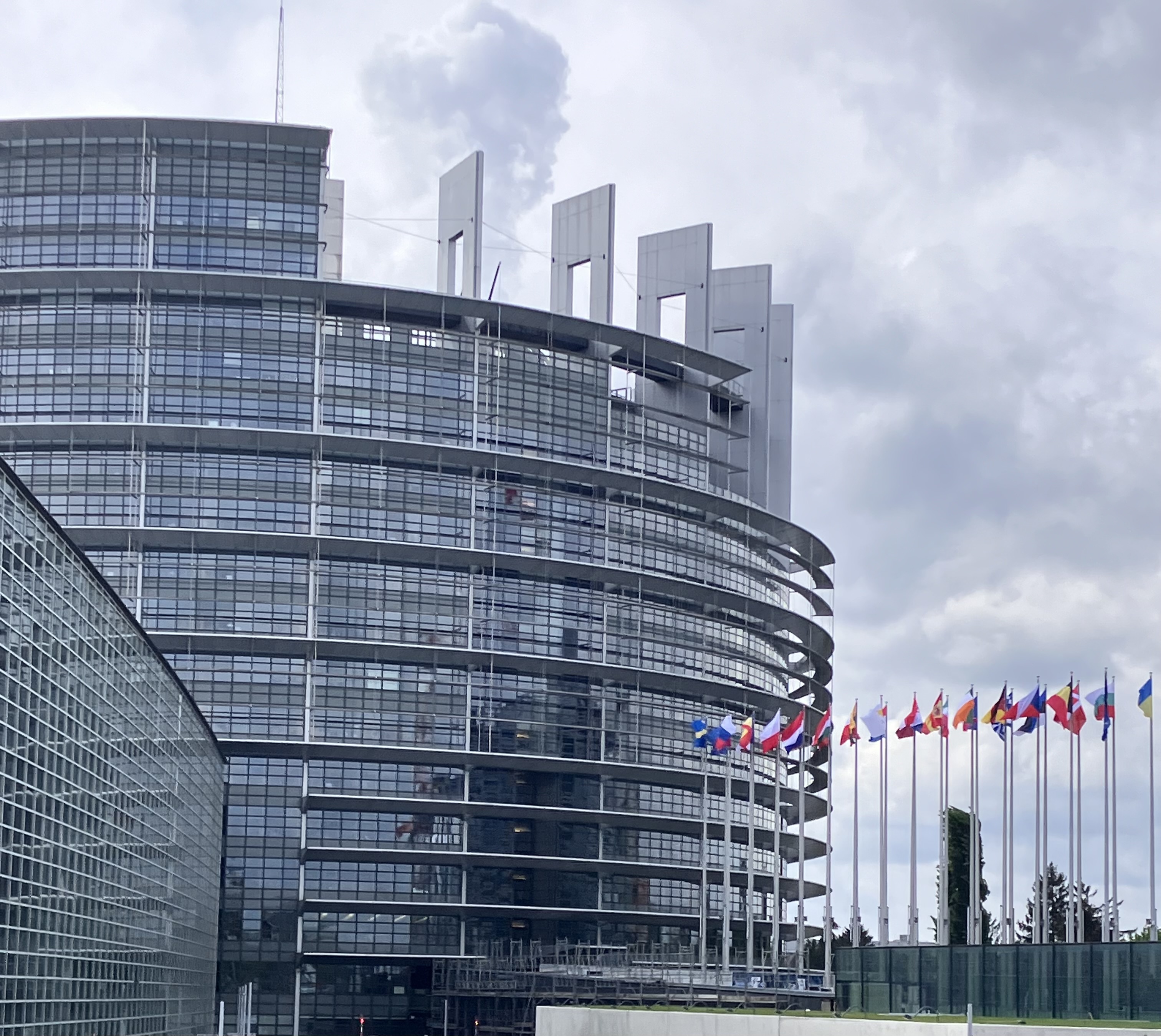 Zu sehen ist das scheinbar unfertige Gebäude des Europaparlaments von außen, sowie die Flaggen der verschiedenen Mitglieder.