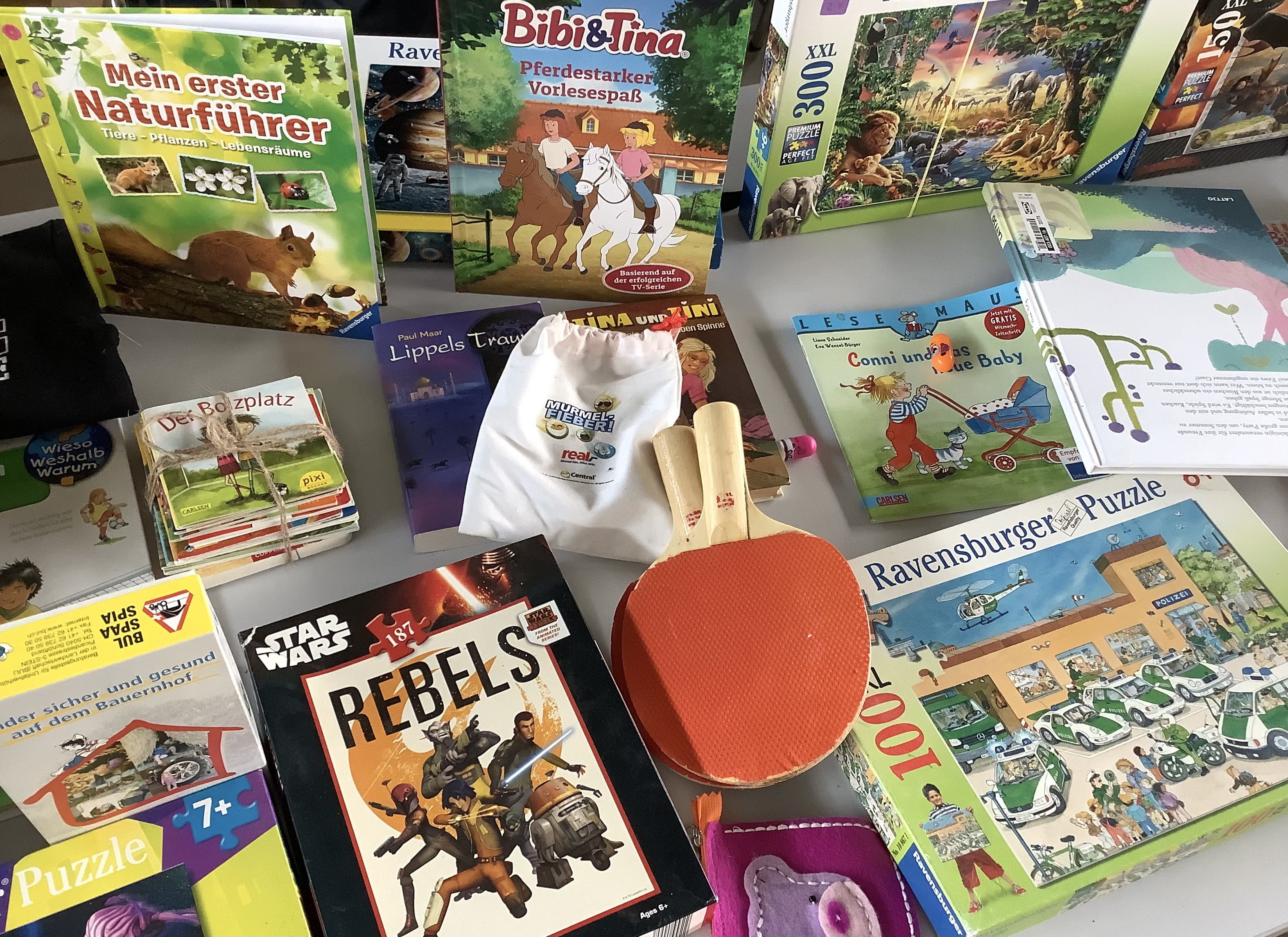 Auf dem Bild sind verschiedene Gegenstände eines Flohmarktstandes abgebildet, wie Puzzles, Kinderbücher oder Tischtennisschläger. 