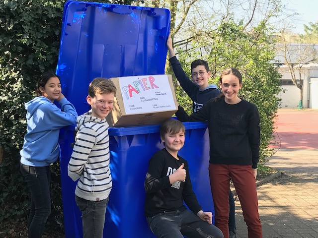 Schüler*innen der Umwelt-AG werfen Altpapier in den großen blauen Altpapier-Container auf dem Schulhof
