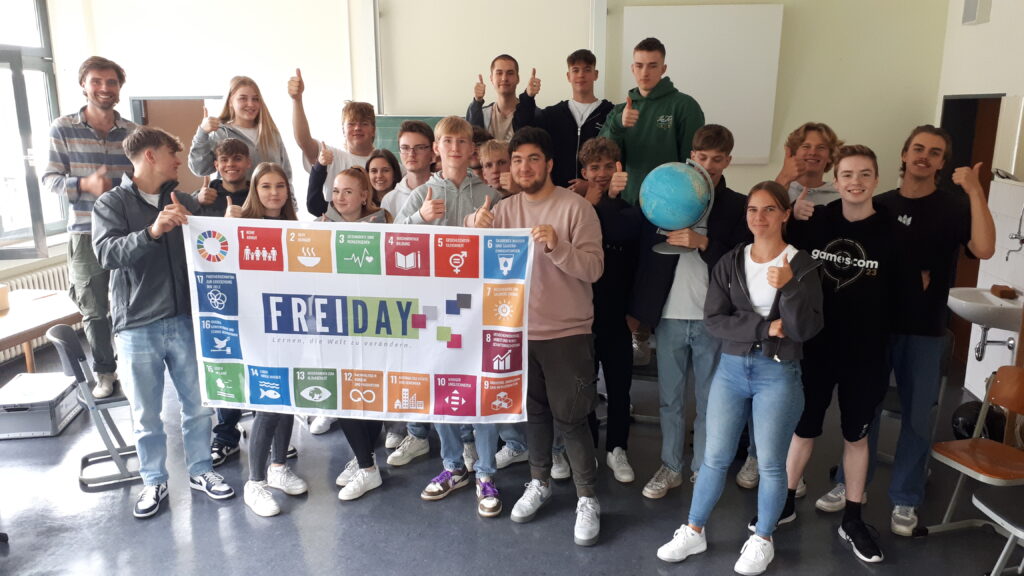 Schüler:innen des Geographie-Leistungskurses posieren in einem Kursraum lächelnd hinter einer erhobenen Flagge mit den aufgedruckten Nachhaltigkeitszielen der Vereinten Nationen.