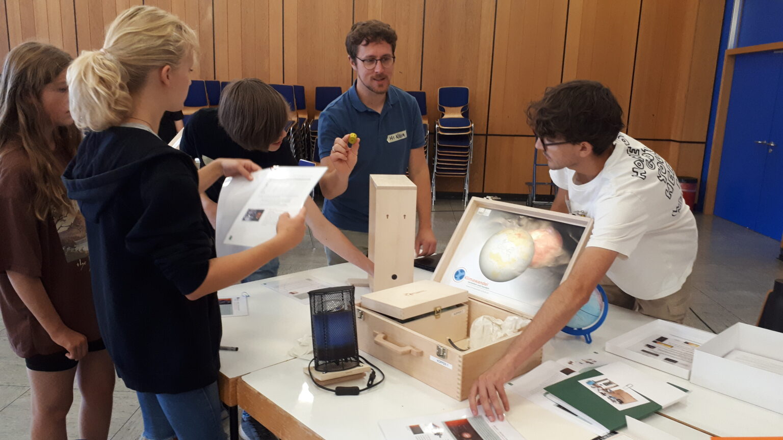 Schüler*innen und eine Lehrkraft experimentieren mit dem auf einem Tisch aufgebauten Klimakoffer