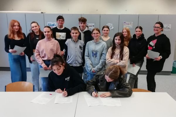 Zwei Schüler*innen unterzeichnen im Bildvordergrund einen Kooperationsvertrag, während im Hintergrund weitere Mitglieder der Öko-E und eine Lehrkraft mit stolzen Gesichtern für das Foto posieren