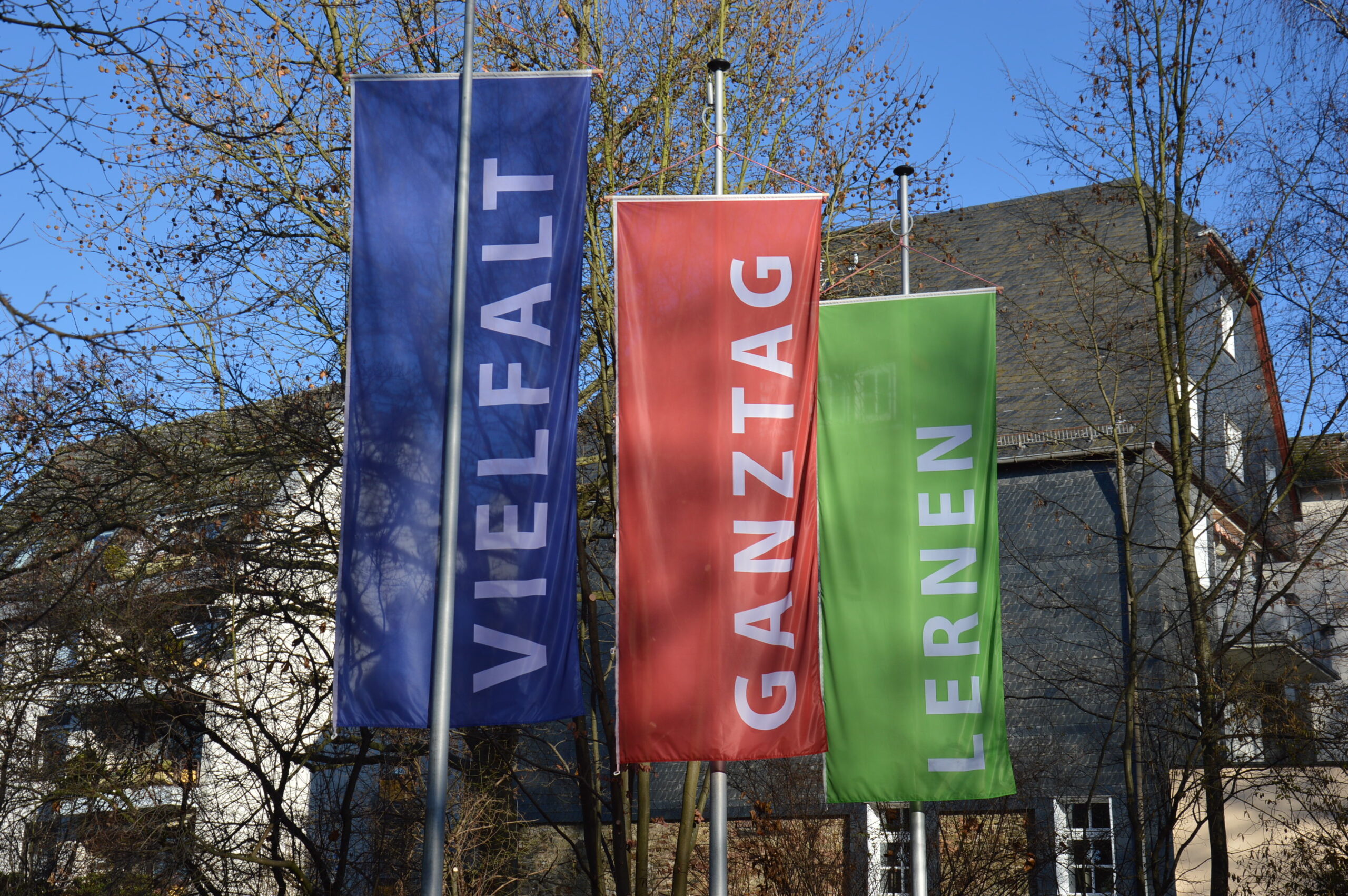 Zu sehen sind die drei Flaggen, die die Schlagworte des Schulmottos der ROS tragen: Vielfalt (blau), Ganztag (rot) und Lernen (grün).