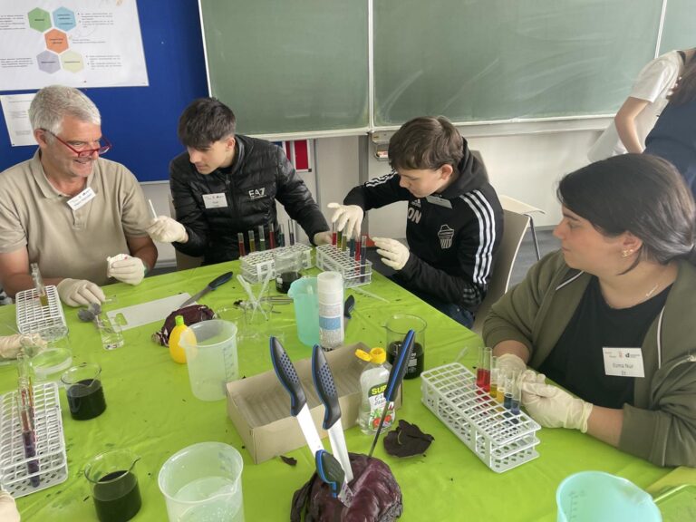 Drei Schüler*innen der ROS untersuchen gemeinsam mit Dr. Björn Hendel mehrere verschiedene Reagenzien an einem Labortisch.