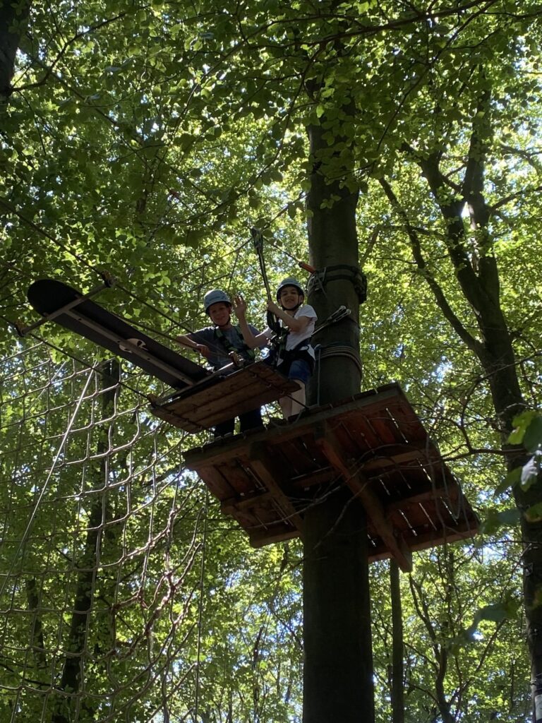 Zwei Schüler der ROS stehen auf einer Plattform des Kletterwalds und machen sich bereit, den weiten Abstand zum nächsten Baum auf einem zwischen zwei Stahlseilen eingehängten Skateboard zu überbrücken.