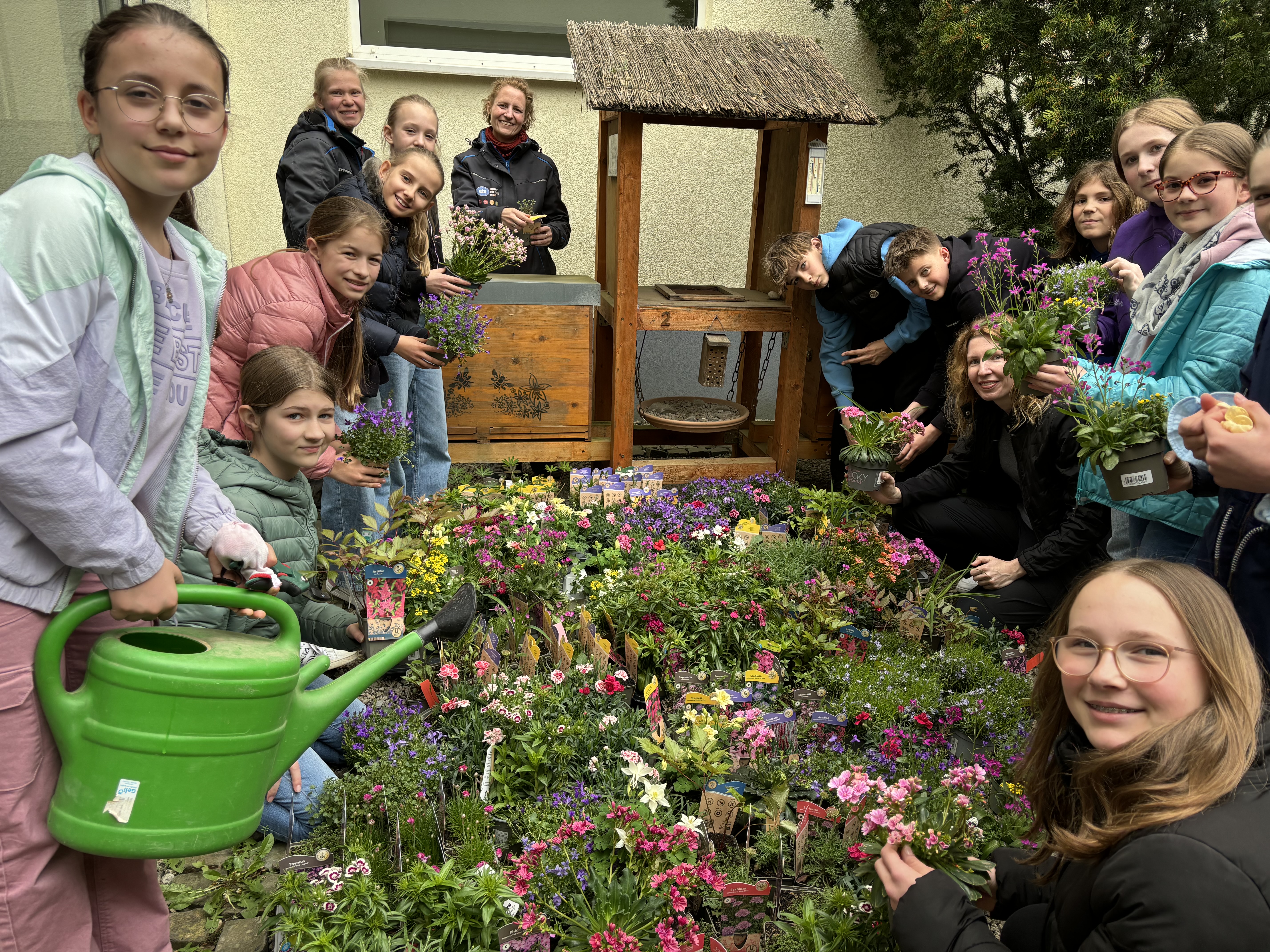 Das Bild zeigt Schülerinnen und Schüler inmitten von den zu pflanzenden Blumen in einem Außenbereich der Schule.