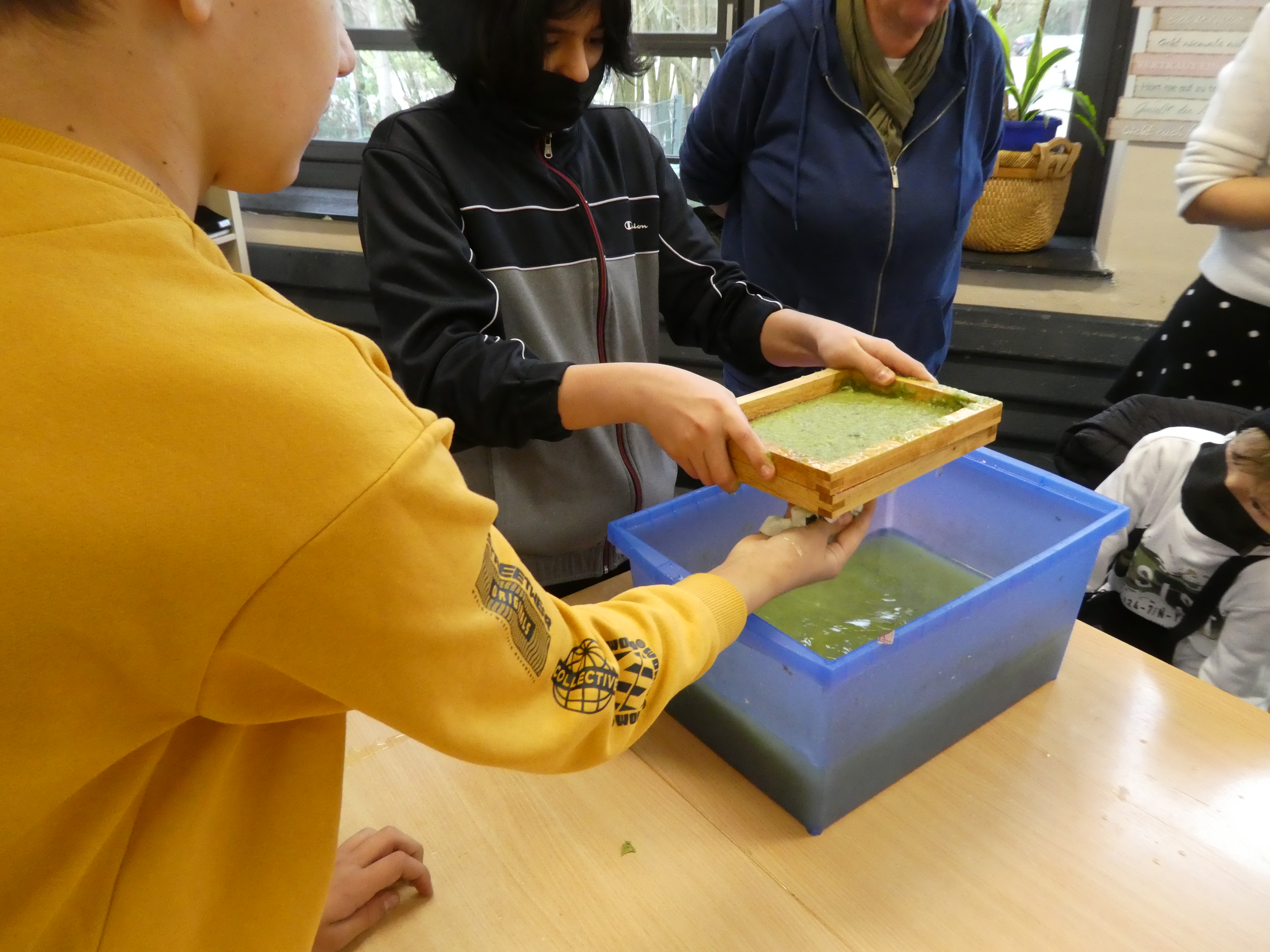 Ein Schüler hat mit einem Rahmen aus einer Wanne eine grünliche Papiermasse geschöpft. Sein Mitschüler tupft mit einem Tuch  überschüssiges Wasser vom Rahmen.
