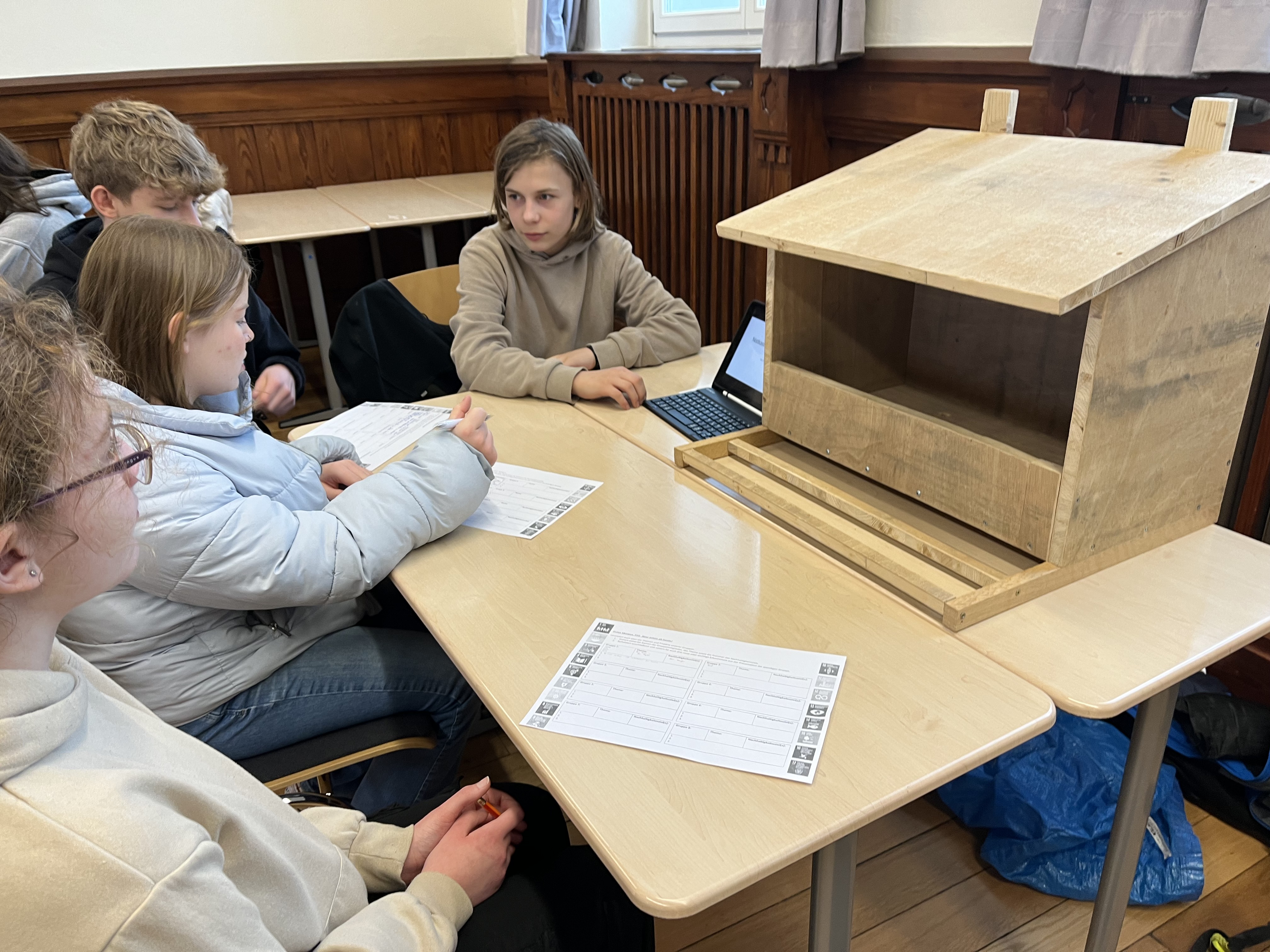 Zu sehen ist der selbstgebaute Nistkasten für Turmfalken aus Holz in einer Größe von etwa 60x60x70cm (BLH) mit seinem Erbauer Jona Teutenberg, der mit Hilfe eine Tablets und einer PowerpointPräsentation einen Vortrag vor 3 SchülerInnen an einem Tisch hält