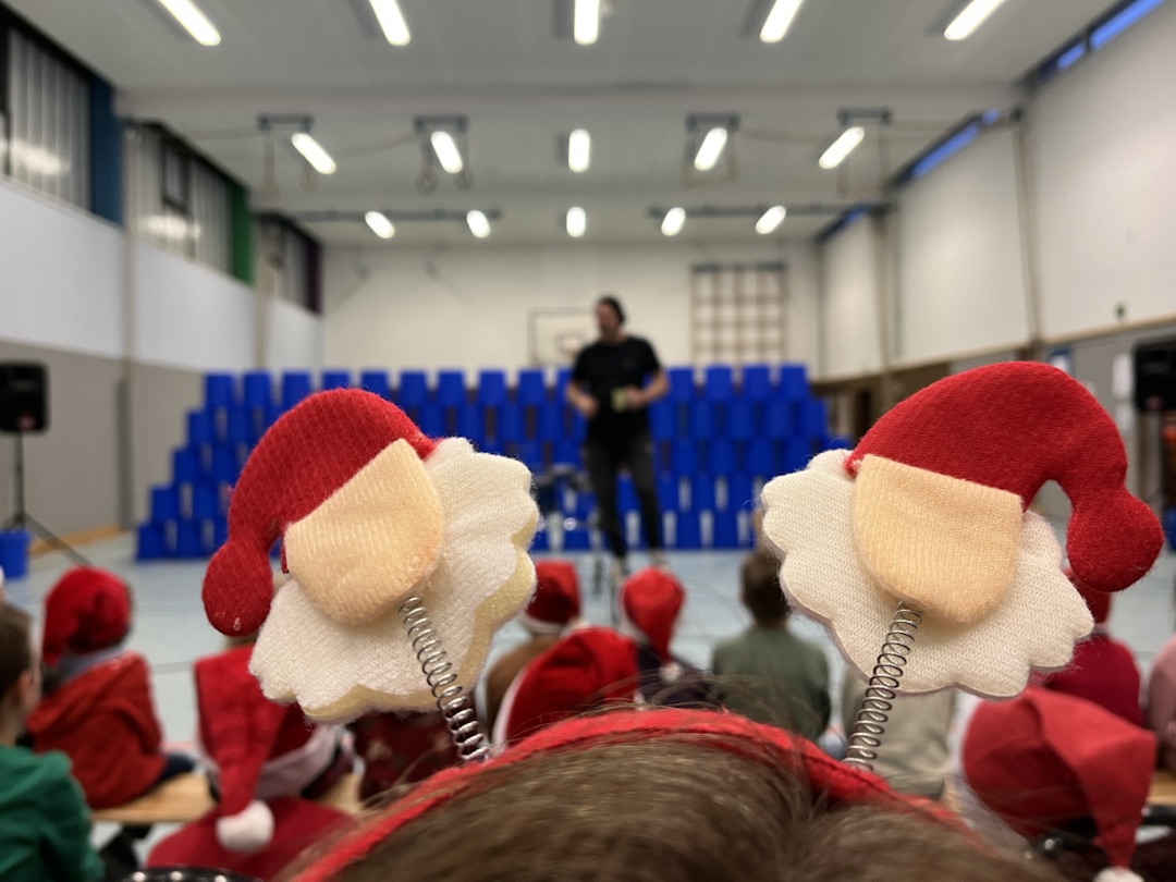 Im Vordergrund ist der Stirnreif eines Kindes mit weihnachtlichen Motiven zu erkennen. Im Hintergrund steht ein Lehrer der Rockschule sowie eine Menge blauer Tonnen, die im weiteren Verlauf als Trommel dienen.