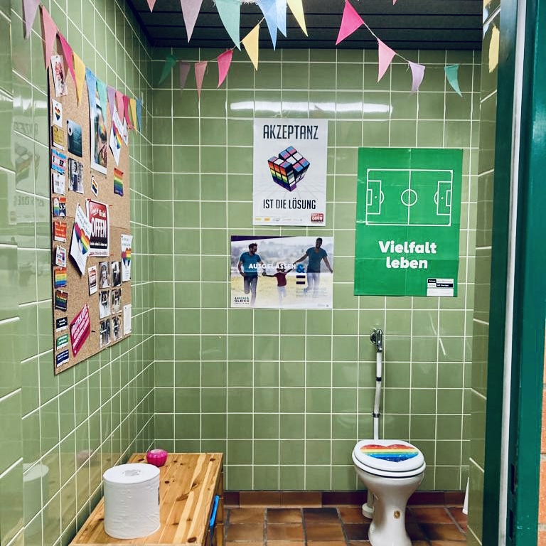 Man sieht eine grün gekachelte Toilette, die mit Wimpelketten und Postern mit Sprüchen zur Vielfalt dekoriert. Auf dem Klodeckel klebt ein Herz in Regenbogenfarben.
