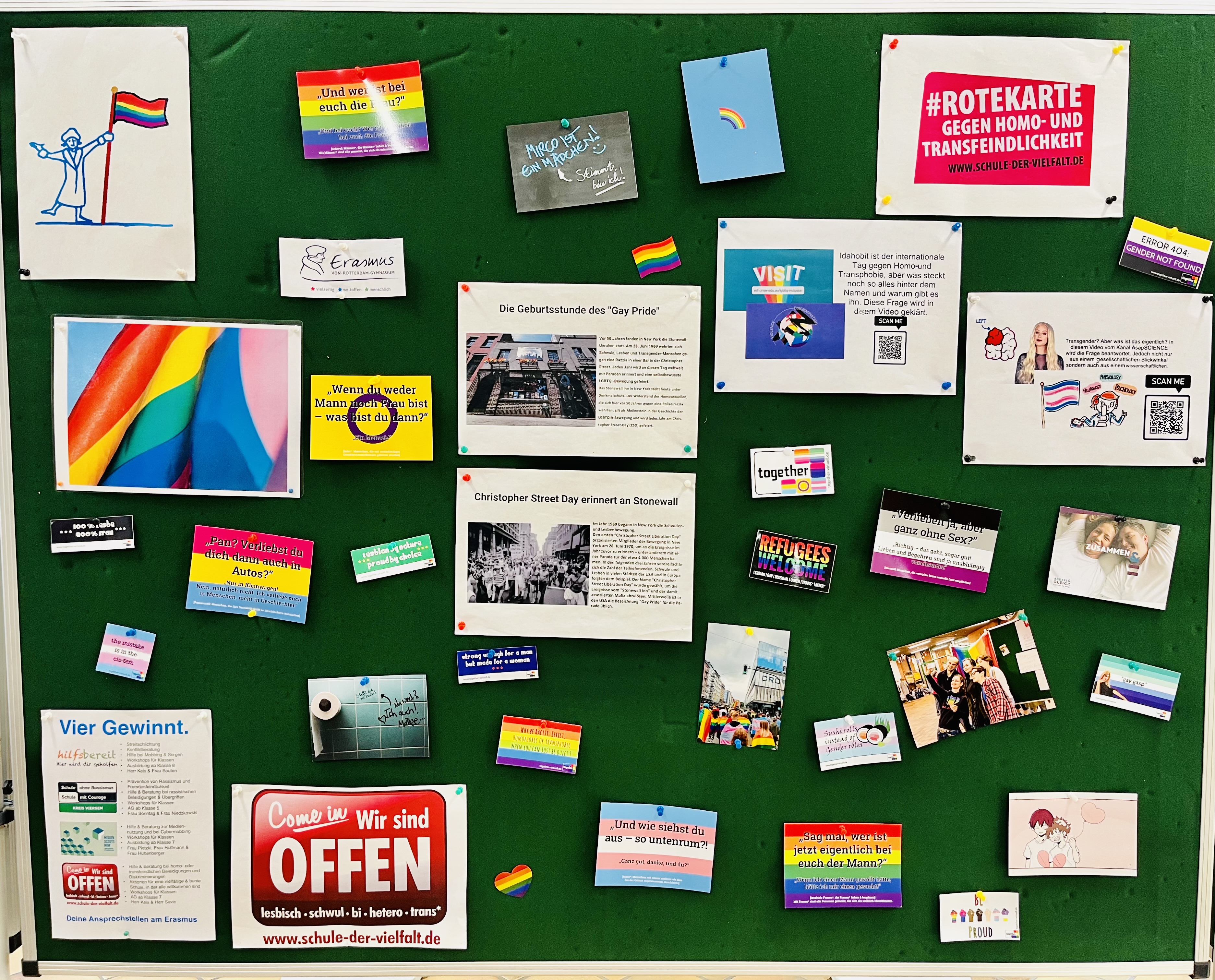 An einer grünen Stellwand hängen verschiedene Informationen zur Gay Pride und zum IDAHOBIT. Zusätzlich sind Regenbogenfahnen zu sehen. 