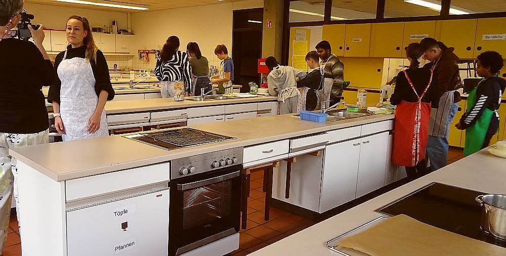 Die Lehrerin beantwortet in der Lehrküche der Schule Fragen vor laufender Kamera. Im Hintergrund arbeiten Schüler und Schülerinnen zwischen den Küchenzeilen an der Zubereitung ihrer Rezepte. 