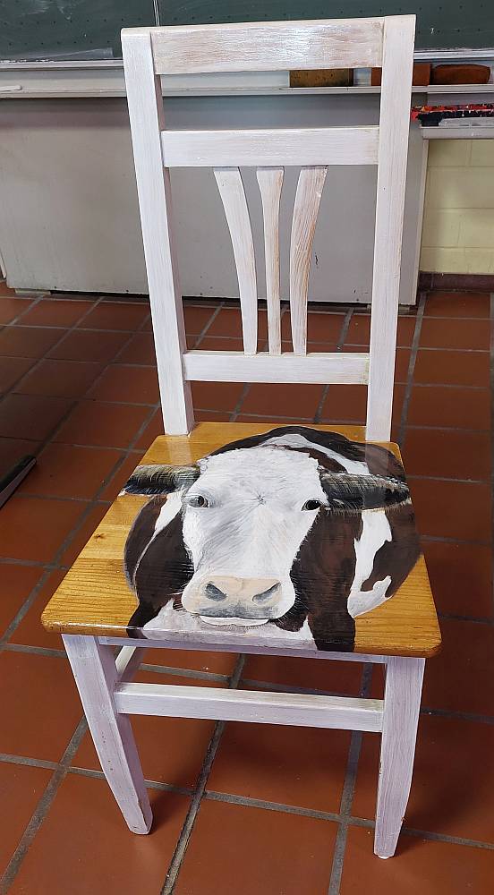Das Bild zeigt einen alten Holzstuhl vom Sperrmüll, der durch einen nachhaltigen, weißen Farbanstrich und dem auf der Sitzfläche aufgebrachten Bild einer Kuh einen neuen Wert erhielt.  