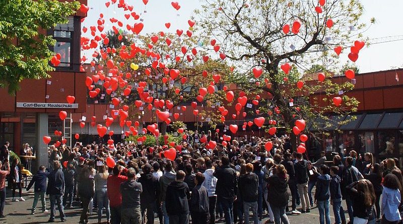 Das Foto zeigt eine große Gruppe von Schülern und Schülerinnen mit roten Luftballons in Herzform vor dem Schulgebäude. 