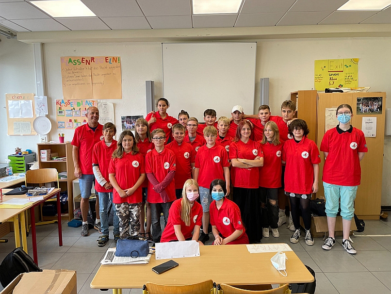Abgebildet ist der Schulsanitätsdienst der Gustav-Heinemann-Schule. Zu sehen ist eine ca. 20 Personen starke Gruppe, bestehend aus Schüler*innen und Lehrer*innen, mit roten T-Shirts.